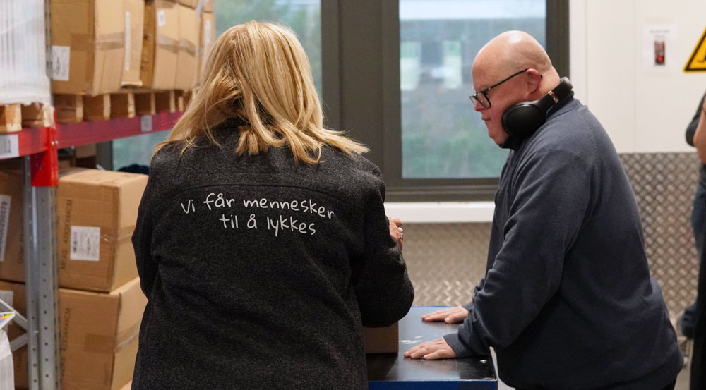 To ansatte i den vernede bedriften Fossheim pakker esker på et arbeidsbord i et lager. En kvinne står med ryggen til med teksten «Vi får mennesker til å lykkes» på jakken, mens Hans Henrik står på motsatt side av bordet og arbeider med pakkingen.