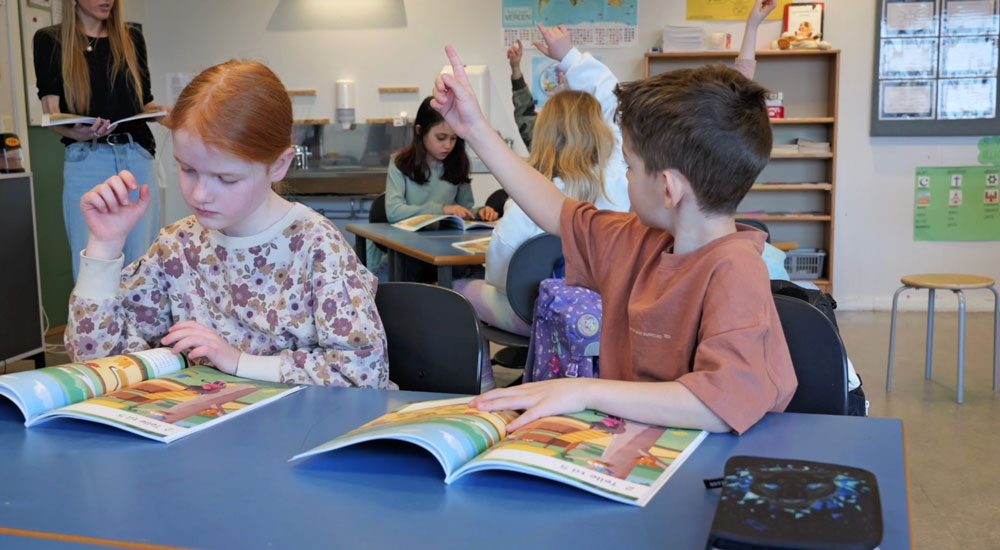 Barn i klasserommet. Alle har arbeidsbøkene åpnet på samtalebildet. Flere elever rekker opp hånden.