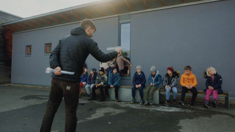 MArius med barn utendørs. Barna sitter på nen benk og lytter til Marius som snakker til dem.