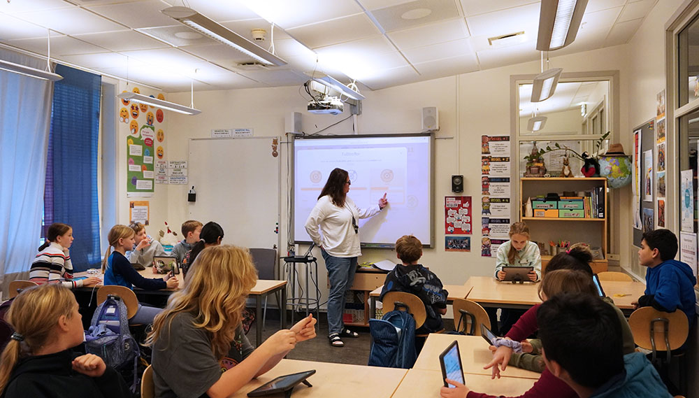 Klasserom med elever. Elevene har datamaskiner og kikker mot en kvinnelig lærer som ser og peker på smart-tavle med bil de fra Mattelabb